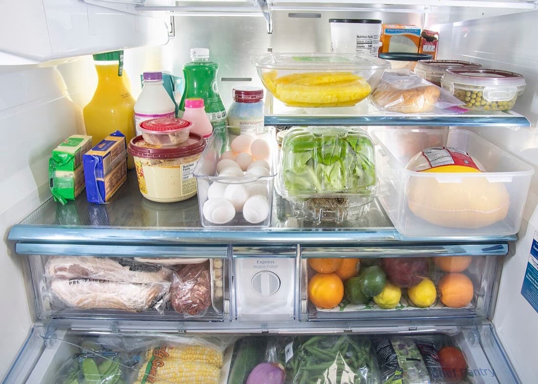 An open fridge: orange juice, milk and a green bottle on the top shelf, butter, hummus, eggs, a salad box and cheese below, then drawers of bacon and citrus, and corn, green beans and snap peas at the bottom.